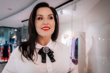 Siyah saçlı ve kırmızı rujlu beyaz bir kadın bir moda mağazasında duruyor, arkasında broş ve giysi vitrinleri olan beyaz bir bluz sergiliyor.