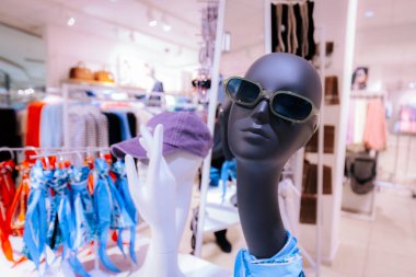 Siyah manken güneş gözlüğü ve mavi bir eşarp, renkli kıyafetler ve aksesuarlarla çevrili bir perakende mağazasında sergileniyor.