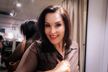 Kafkas kadın, uzun siyah saçlı, kahverengi bir çantayla bir giyim mağazasında poz veriyor, arka planda aynalar ve elbise askılarıyla çevrili.