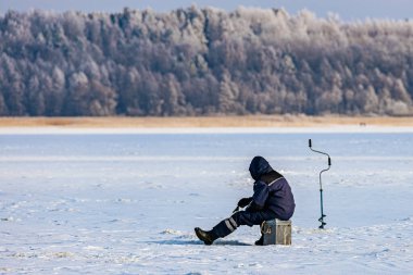 Kışın giyinmiş bir insan, donmuş bir gölde buzda balık tutarken ve arka planda karla kaplı bir orman varken bir kutunun üzerinde oturur.