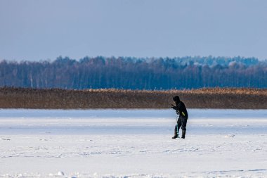 Burtnieks, Letonya - 09 Şubat 2026: Siyah kış giysileri giyen kişi karla kaplı bir alanda dikiliyor, uzaktaki ağaçların olduğu bir akıllı telefonu kontrol ediyor