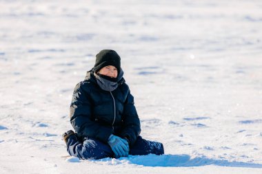 Siyah kışlık paltolu ve eldivenli çocuk karla kaplı bir yüzeyde oturuyor, parlak, güneşli bir kış manzarasıyla çevrili.