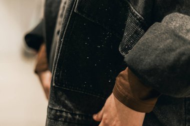 Koyu renk kot ceketli, kahverengi gömlekli, elleri cepli, kumaşın dokusunu yumuşak, nötr bir arka plana doğru çizen birinin yakın görüntüsü.