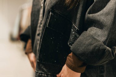 Koyu renk kot ceketli, cebi olan bir şahsın yakından görüntüsü, modern bir giyim mağazasının arka planına karşı kurulmuş, bulanık bir giysi.