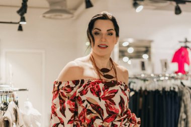 Çiçekçi elbiseli, geniş beden bir kadın elbise mağazasında dikiliyor. Etrafı giysi rafları ve aydınlatmalarla çevrili.