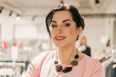 Beyaz kadın bir giyim mağazasında pembe bir bluz ve kalın bir kolye giyerek dikiliyor. Arka planda elbise askıları ve diğer müşteriler görünüyor.