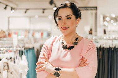 Beyaz kadın bir giyim mağazasının içinde kolları çapraz duruyor, pembe bir elbise ve mücevher sergiliyor, arka planda elbise askıları var.