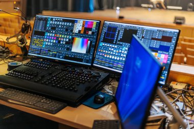 İkili monitörler, çalışma alanı etrafında çeşitli kablolar ve ekipmanlarla bir performans mekanındaki ahşap bir masada ışıklandırma kontrol yazılımını gösteriyor.