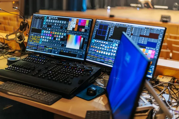 İkili monitörler, çalışma alanı etrafında çeşitli kablolar ve ekipmanlarla bir performans mekanındaki ahşap bir masada ışıklandırma kontrol yazılımını gösteriyor.