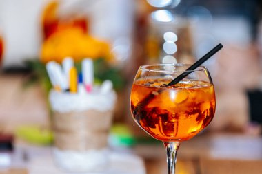 Valmiera, Letonya - Mart 06, 2026: Buzlu bir bardakta portakal kokteyli ve ahşap bir masada siyah bir pipet, arka planda renkli bulanık süslemeler ve kırtasiye malzemeleri