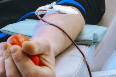 Erkek kan donörü elinde bir stres topu tutarken kan bağışı yapıyor. Kolunda serum ve bandaj var.
