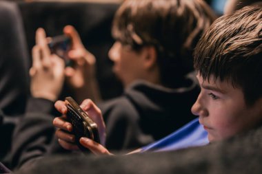 İki genç çocuk birbirine yakın otururken akıllı telefon kullanıyor. Hafif ışıklandırma iç mekan düzenini güçlendiriyor.