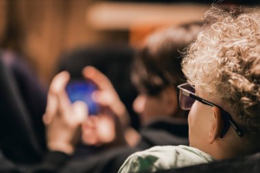 İki çocuk kapalı alanda rahat bir ortamda akıllı telefonlarına odaklanırken, bir çocuk gözlük takıyor ve kıvırcık saçlı, arka plan ise yavaşça bulanık