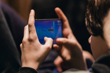 Çocuk iki eliyle akıllı telefon kullanıyor, ekranda canlı grafiklerle meşgul, kapalı arka planda rahat bir şekilde oturuyor.