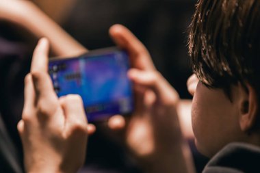 Genç bir çocuk canlı oyun grafikleri gösteren akıllı bir telefonla meşgul, kapalı bir alanda, bulanık bir çevrede oturuyor.