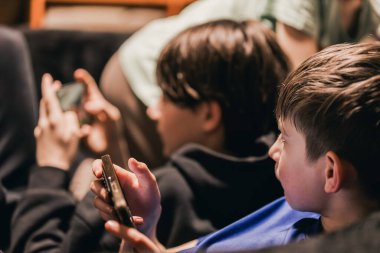 İki çocuk koltukta otururken akıllı telefon kullanıyor, biri siyah kapüşonlu, diğeri mavi tişörtlü, hafif ışıklandırma sahneyi büyütüyor.