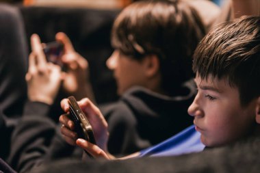 İki çocuk kanepede otururken akıllı telefon kullanıyor, bir çocuk cihazına odaklanıyor, rahat bir atmosfer yaratıyor.