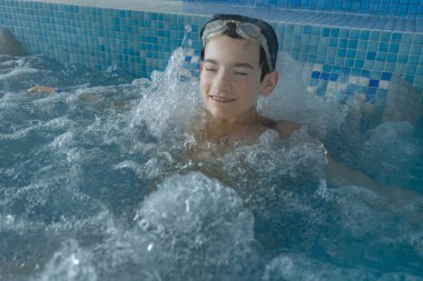 Yüzme gözlüğü takan genç bir çocuk, etrafında mavi mozaik fayanslar ve su sıçrayan köpüklü bir jakuzide rahatlatıcı bir anın tadını çıkarıyor.