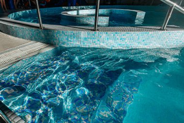 Mozaik kiremit desenli dairesel havuz ve berrak mavi su, spiral bir tasarım ve güneş ışığıyla aydınlatılan paslanmaz çelik korkuluklar.