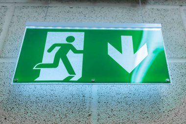 Yeşil acil çıkış levhasında koşan bir figür ve aşağı doğru eğilen bir ok var, aydınlanmış ve dokulu bir duvara monte edilmiş, bireyleri güvenli bir yere götürüyor.