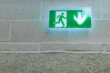 Yeşil acil çıkış levhasında koşan bir figür ve aşağıya bakan bir ok var. Kamu binası koridorundaki beton bir duvara yerleştirilmiş.