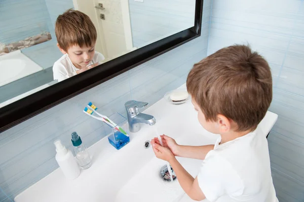 Una persona poco caucásico sano lindo niño en blanco camisa lavándose las manos bajo agua dulce en lavabo en azul cuarto de baño en casa — Foto de Stock