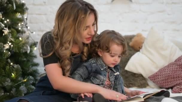 Felice giovane famiglia caucasica madre femmina e piccola ragazza riccia guardando interessante libro in arredamento natalizio interno — Video Stock