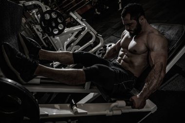 Güçlü, genç, sakallı bir atlet. Egzersiz makinesinde bacak presi yaparken ağır ağır oturuyor.