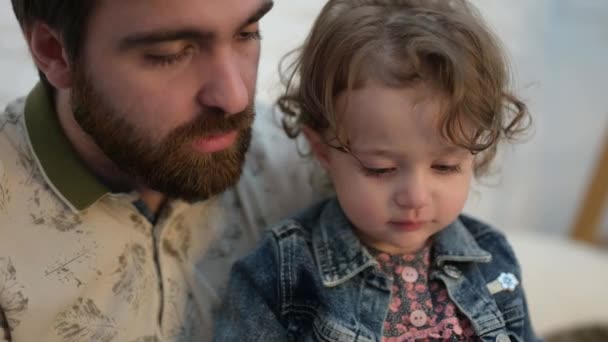 Ung skäggig man med skägg titta på bok med liten dotter söt lockig flicka inomhus — Stockvideo