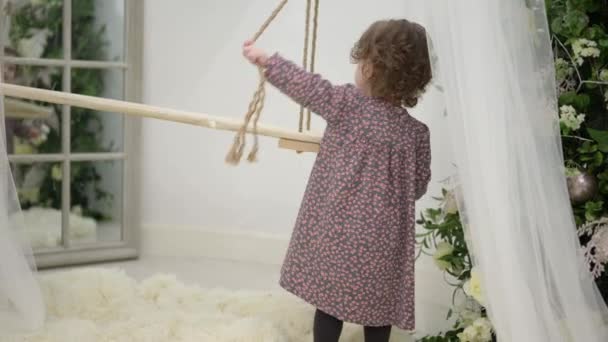 在白色工作室里，穿着连衣裙的卷曲小女孩玩木绳秋千的生活方式休闲活动 — 图库视频影像