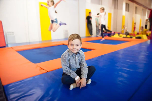 Petit garçon en bonne santé assis sur le trampoline parmi les enfants sautant à l'intérieur dans le club d'activités de loisirs — Photo