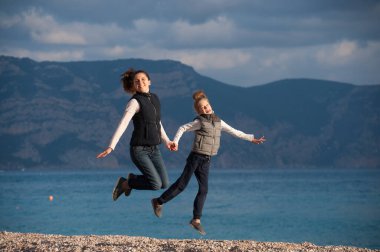 happy active mother and child in sleeveless jackets jumping on sea beach on blue yellow sunset clipart