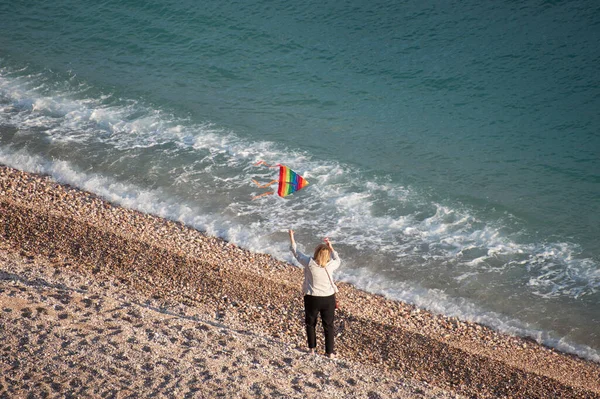 Γυναίκα ηλικία χειραφέτησης έννοια της παλιάς γυναίκας με φέρουν πολύχρωμο χαρταετό στο ηλιοβασίλεμα όμορφη παραλία της θάλασσας με τα κύματα του νερού και φυσώντας αέρα την άνοιξη — Φωτογραφία Αρχείου