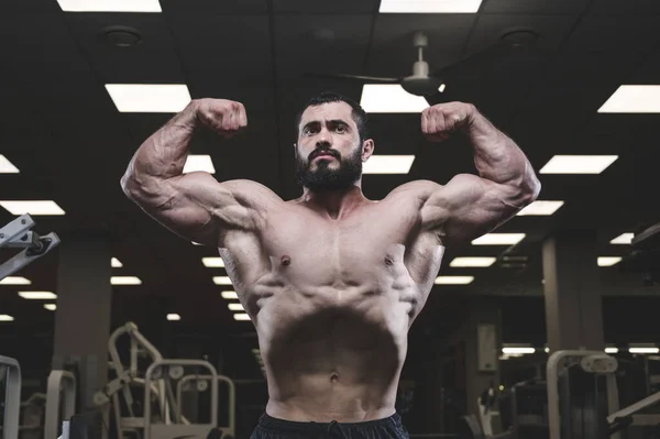 Musculoso forte jovem caucasiano com barba mostrando poderoso bíceps braços músculo posando no ginásio fitness escuro — Fotografia de Stock