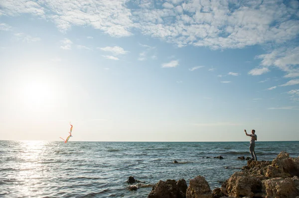 Ένα ενεργό αρσενικό παίζει με ιπτάμενο χαρταετό στέκεται στη θάλασσα βραχώδη ακτή στο ηλιοβασίλεμα δραστηριότητα αναψυχής καλοκαίρι — Φωτογραφία Αρχείου