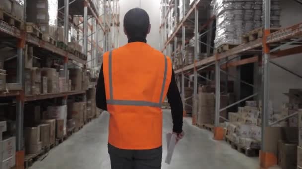 Ein Arbeiter in orangefarbener Weste läuft mit Kisten voller Produkte mit Kamerafahrten am Lager entlang — Stockvideo