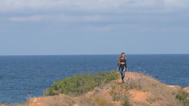 Активная спортивная девушка гуляет с тремя собаками вдоль морского берега с парусной яхтой позади во время досуга в отпуске — стоковое видео