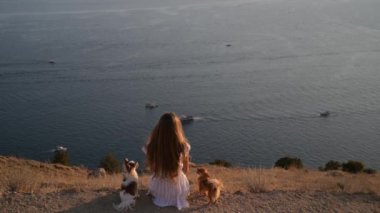 Yaz günbatımı meditasyonu sırasında denizde yelkenli tekneli iki küçük chihuahua köpeğiyle deniz kıyısında oturup hayal kuran güzel bir genç kadın manzarası.
