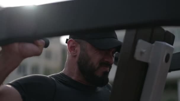 Silný mladý vousatý sportovec muž v černé čepici a roztržené tričko zvedání těžké váhy na cvičení cvičební stroj — Stock video