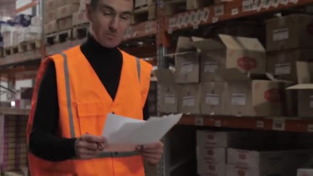 Человек работник в оранжевом жилете прогуливаясь по складу проверки документов с коробками продуктов с движения камеры — стоковое видео
