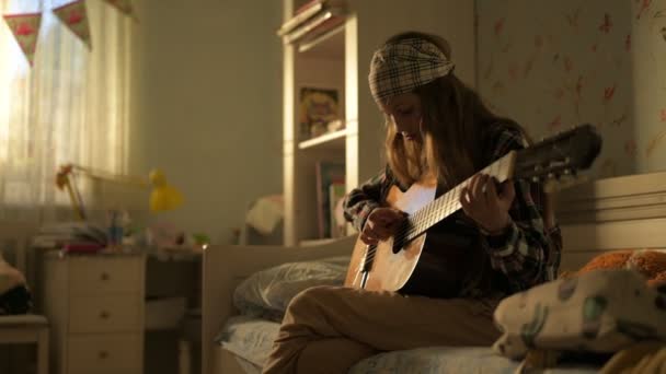 Young teen girl in plaid shirt and rock bandana playing guitar sitting indoors in her room lit with sunset in window — Stock Video