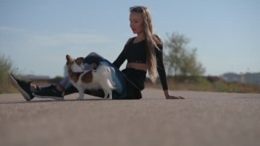 Genç bayan köpek sahibi eğitmeni, yaz sporu aktiviteleri sırasında asfalt yolda boş boş oturarak numaralar yapmak için köpeğini eğitiyor.