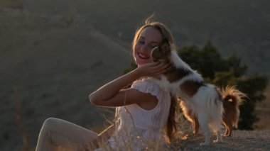 Mutlu, gülümseyen genç bir kadın, Chihuahua köpeklerini okşayarak günbatımı dağında dostluk ve şefkatle omzuna yaslanıyor.