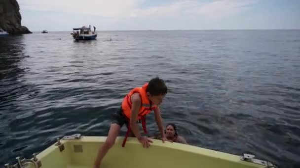 Summer sailing cruise of child in life jacket vest jumping to water to his mother from boat board near rocky coast and sailing boat with people during vacation travel leisure — Stockvideo