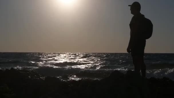 En aktiv hane i mössa och ryggsäck står på havets strand på vacker solnedgång under sommarsemester — Stockvideo