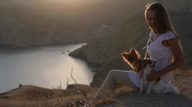 Gün batımında deniz koyunun yanındaki güzel dağda oturan mutlu genç bir kadın sahibi yaz tatilinde yürüyüş yapan iki sevimli köpekle birlikte.