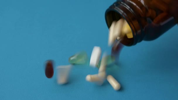 Many tablets pills and capsules of vitamin and medicine falling from glass brown pharmacy bottle on blue surface — Stock Video