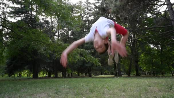 Aktives gesundes kleines Kind in roten Shorts und weißem Trikot, das während der Freizeitaktivität im grünen Park in Zeitlupe Purzelbäume schlägt — Stockvideo