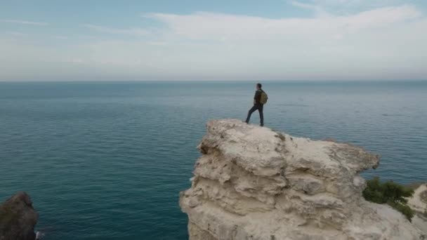 Antenn utsikt skjuten från drönare av en ensam manlig vandrare med ryggsäck på klippkanten nära havet och himmel horisont — Stockvideo