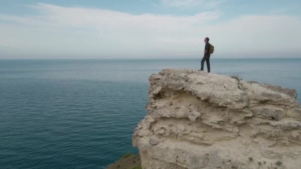 Een persoon mannelijk met rugzak staan op de rand van rotsachtige berg op blauwe hemel en zee horizon achtergrond geschoten door vliegende drone luchtfoto camera beweging — Stockvideo